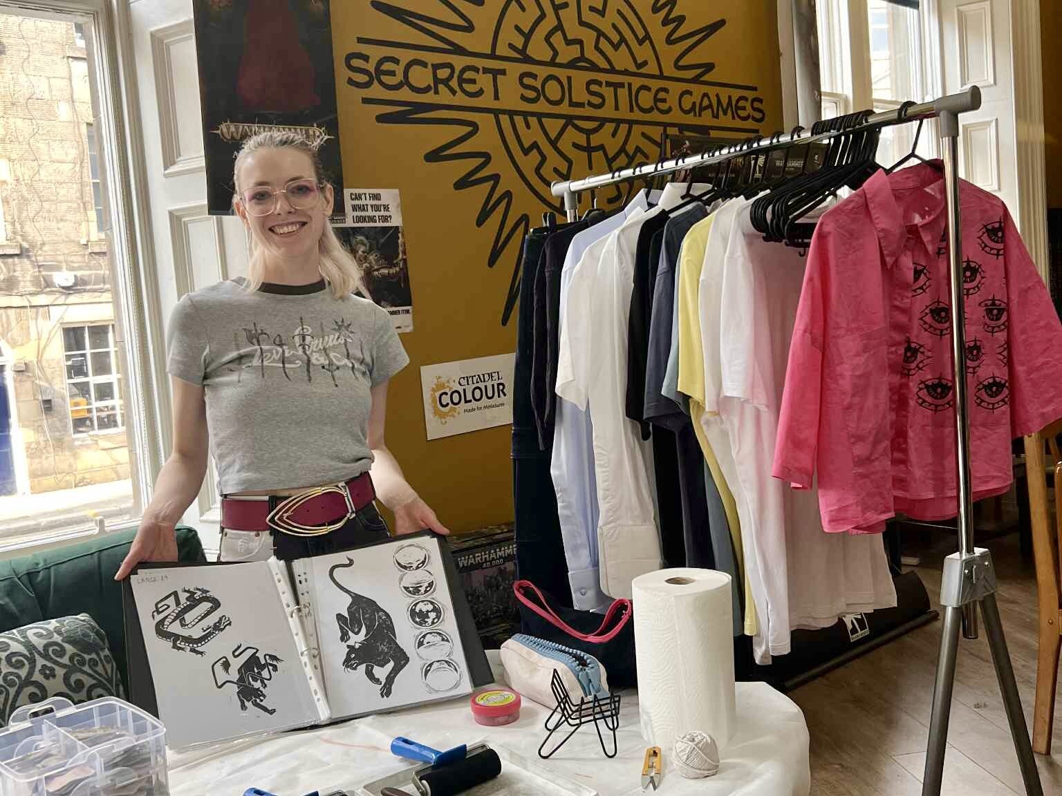 Avery behind a hand-printing stall holding open a book displaying linocut designs, beside a rail of clothing to be printed.
